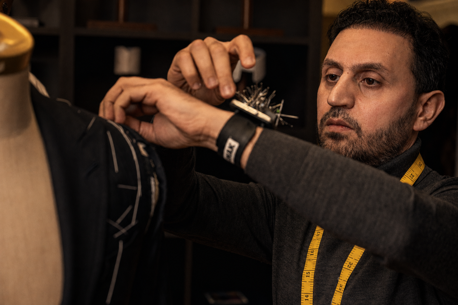 Master tailor with a wrist pin cushion pinning the shoulder of a basted navy jacket on a dress form